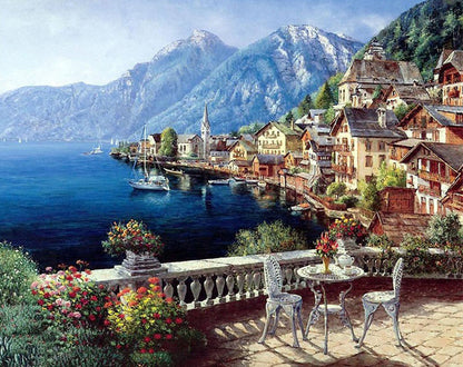 Scenic view of a lakeside village with mountains and a table with chairs.