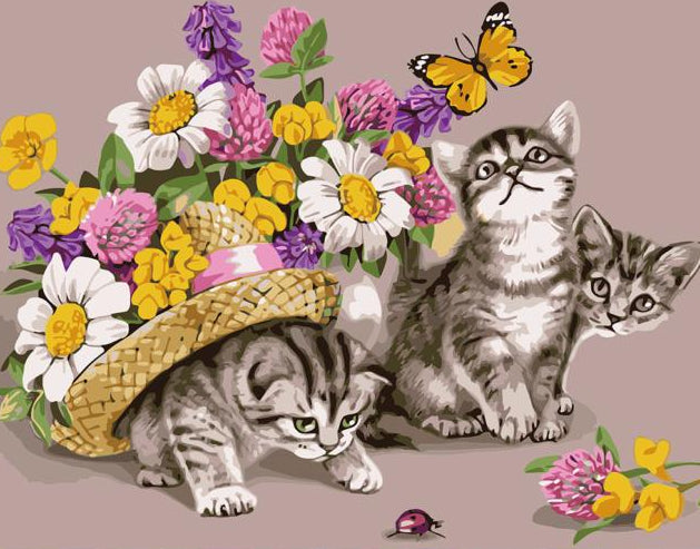 A square diamond painting or craft kit design featuring two tabby kittens, a basket of colorful flowers, and a yellow butterfly on a dusty pink background, displayed on a wooden surface with paint pots, brushes, and a rolled sheet.