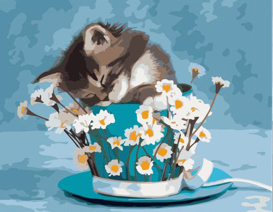 A canvas painting kit showing a brown and white kitten sleeping in a teal cup filled with white daisies, displayed on a wooden surface against a white brick wall. Paint pots, brushes, and a rolled-up canvas are visible in the foreground.